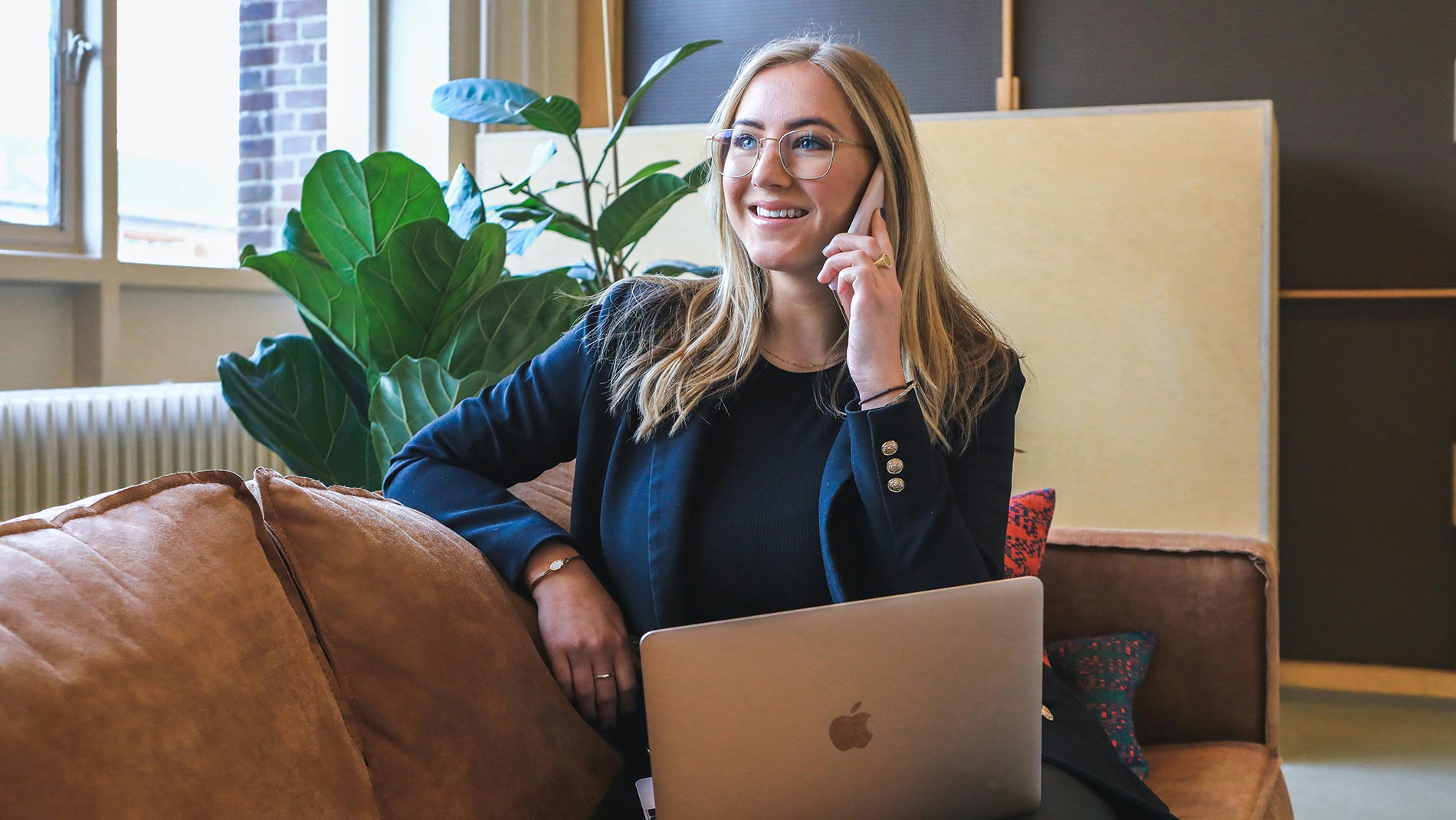Smiling woman on phone with laptop seated on a couch in a modern office space