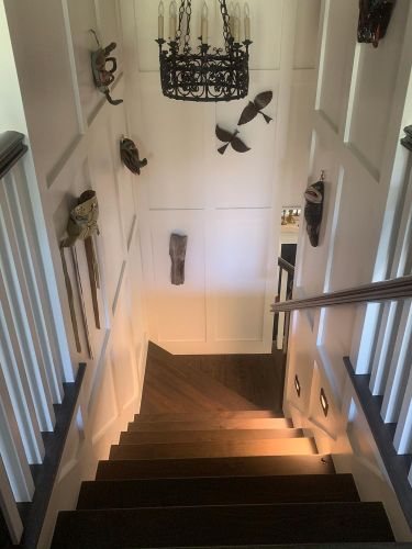 Downward staircase view with ornate chandelier and art masks on white panel walls